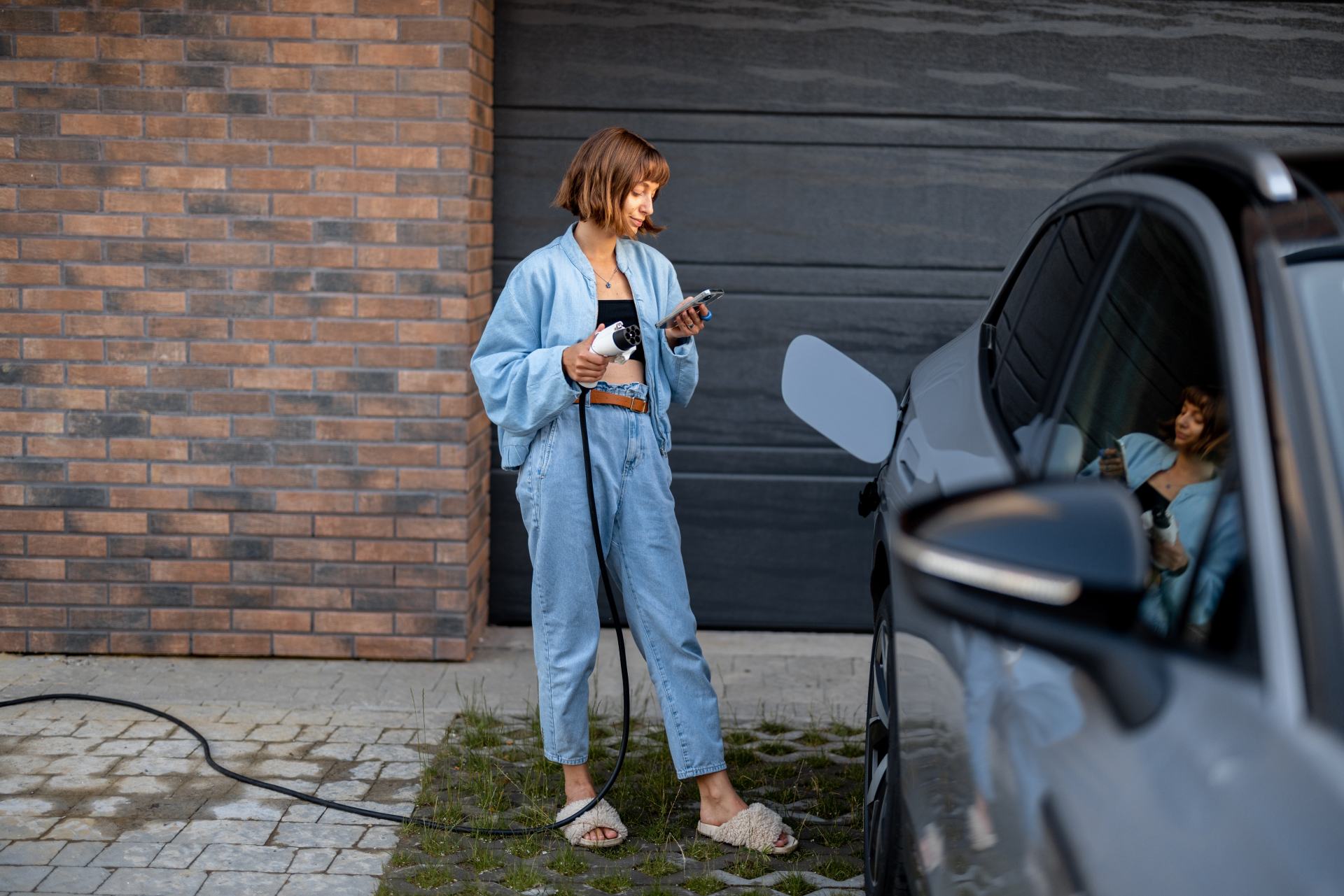 Eine junge Frau steckt die Ladevorrichtung für ihr Auto ein, während sie mit einem Smartphone in der Nähe eines Garagentors steht.