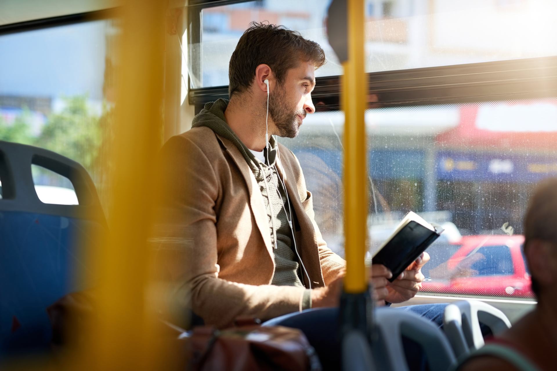 Mann im Bus schaut aus dem Fenster mit Kopfhörern in den Ohren und einem Buch in den Händen.