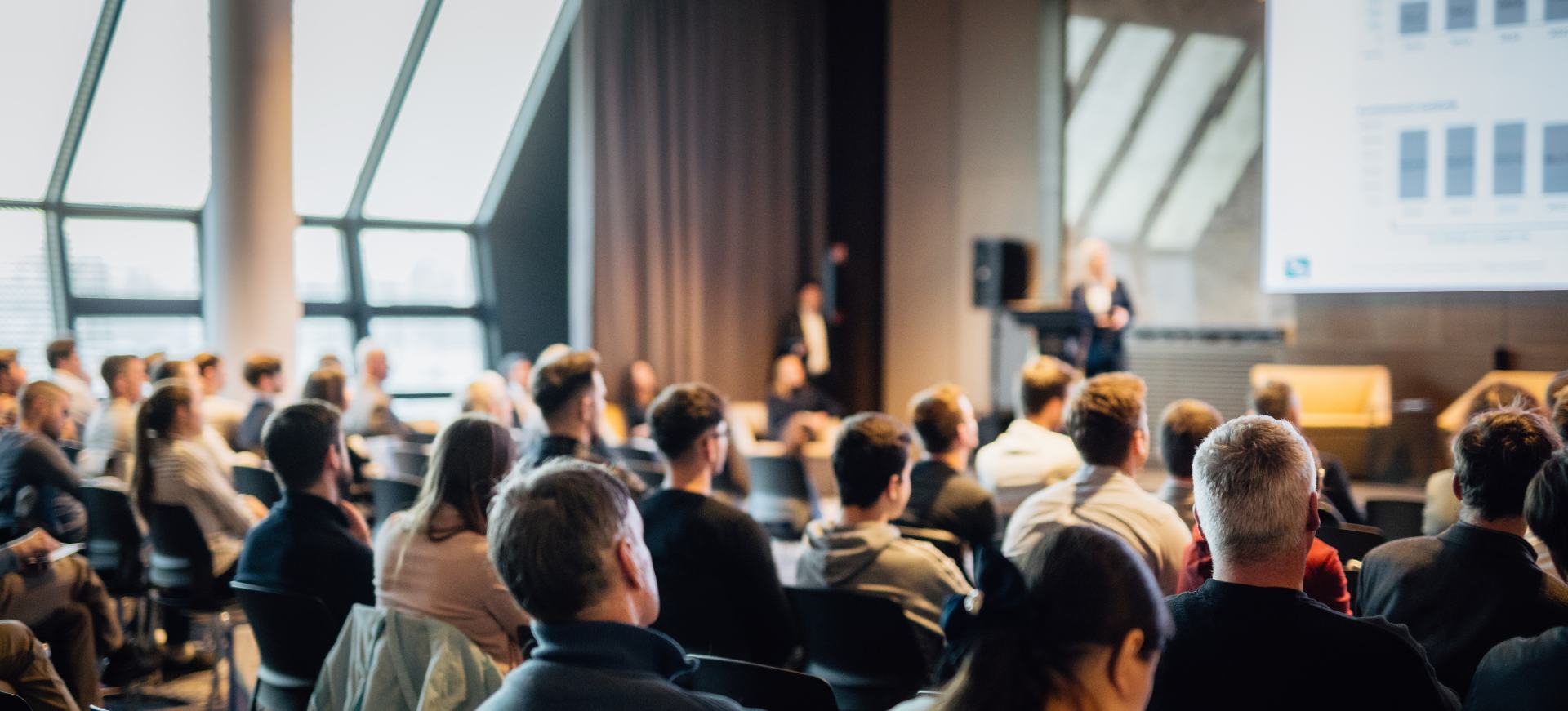 Ein Redner hält einen Vortrag im Konferenzsaal einer Veranstaltung. Rückansicht von nicht erkennbaren Personen im Publikum des Konferenzsaals.